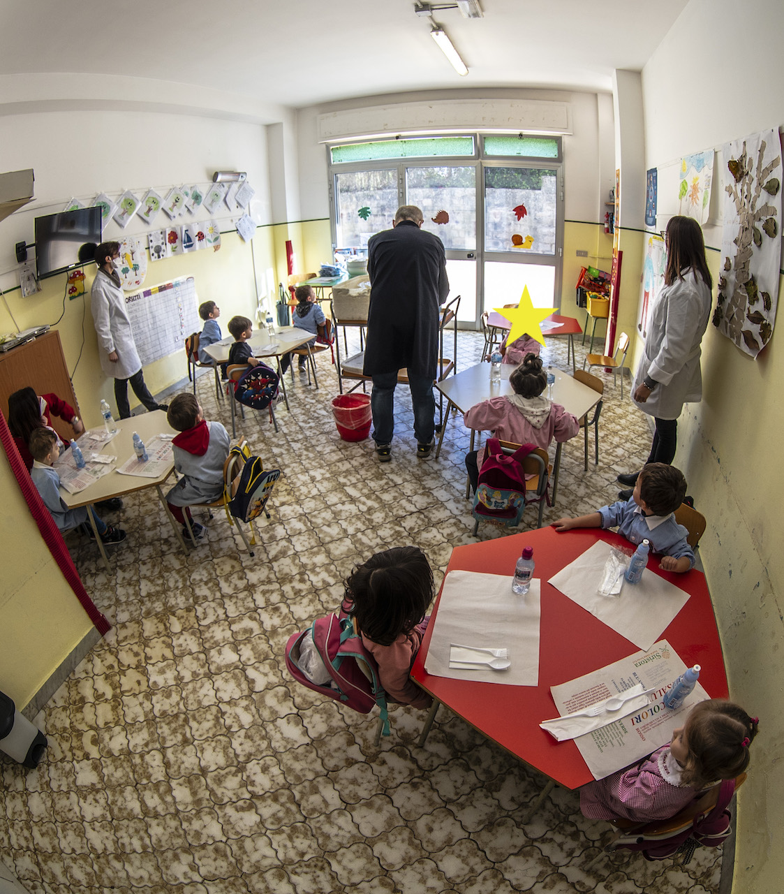 Bambini in aula seduti sui banchetti che attendono il pasto mentre il collaboratore prepara il servizio mensa